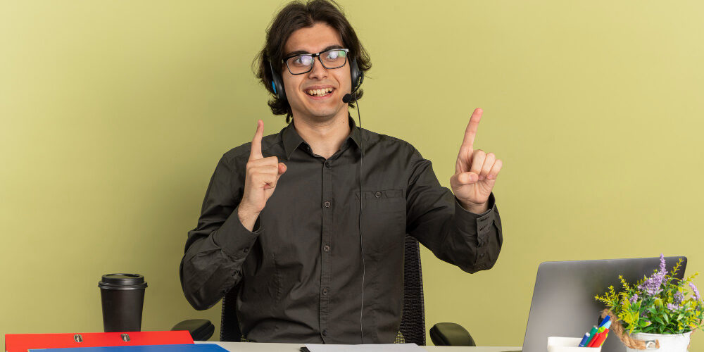 young-smiling-office-worker-man-headphones-optical-glasses-sits-desk-with-office-tools-using-laptop-points-up-isolated-green-background-with-copy-space headsets